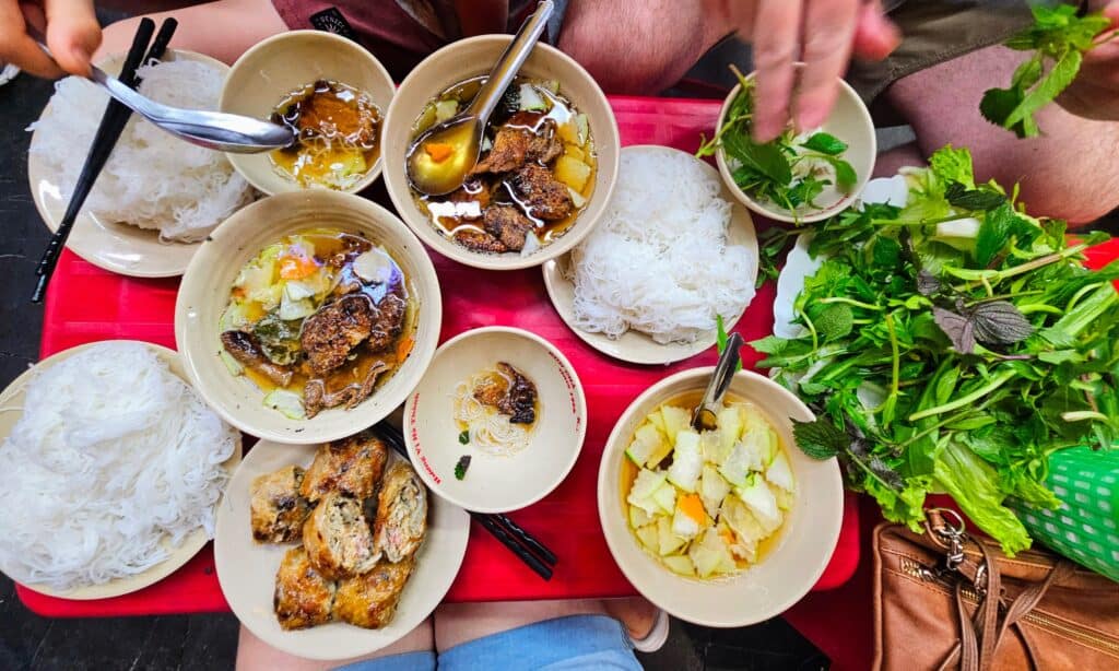 Overhead view of a traditional Vietnamese meal featuring bowls of grilled pork in savory broth, plates of white rice vermicelli, fresh herbs, pickled vegetables, and crispy spring rolls. The colorful spread is laid out on a red table, capturing a vibrant street food experience in Hanoi.