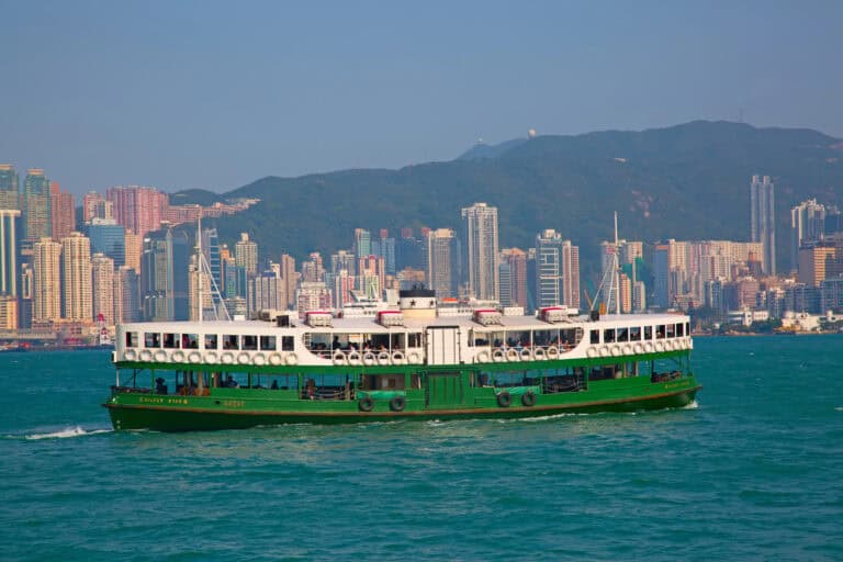A green and white Star Ferry boat sails across Victoria Harbour with the skyline of Kowloon in the background, featuring a dense cluster of high-rise buildings backed by green hills. The iconic ferry connects Hong Kong Island and Kowloon, offering scenic and practical transport between the two areas.