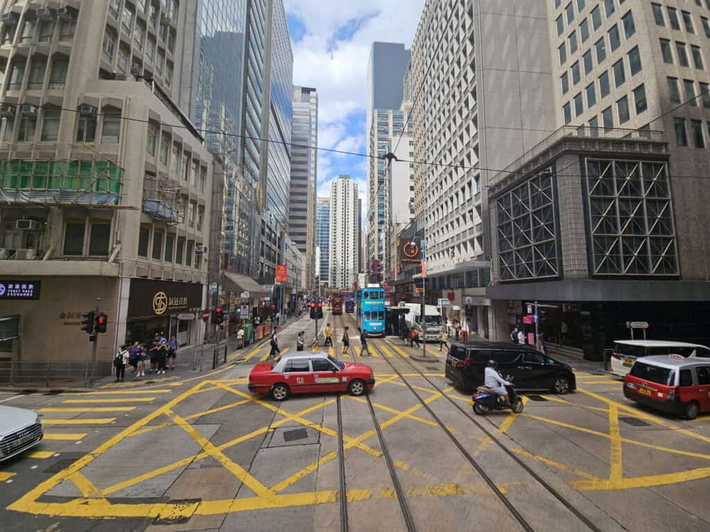 A busy street intersection on Hong Kong Island with red taxis, double-decker trams, and pedestrians crossing under tall office buildings. The energy and vertical density reflect the fast-paced, business-focused vibe of Central.