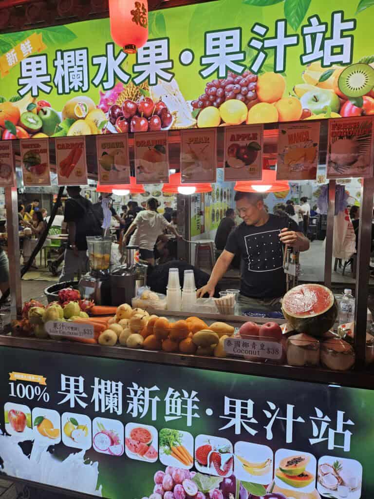 A colorful night market fruit juice stall in Kowloon selling freshly cut tropical fruits and juices under a neon-lit sign in Chinese. The lively market scene emphasizes the local, vibrant character of Kowloon.