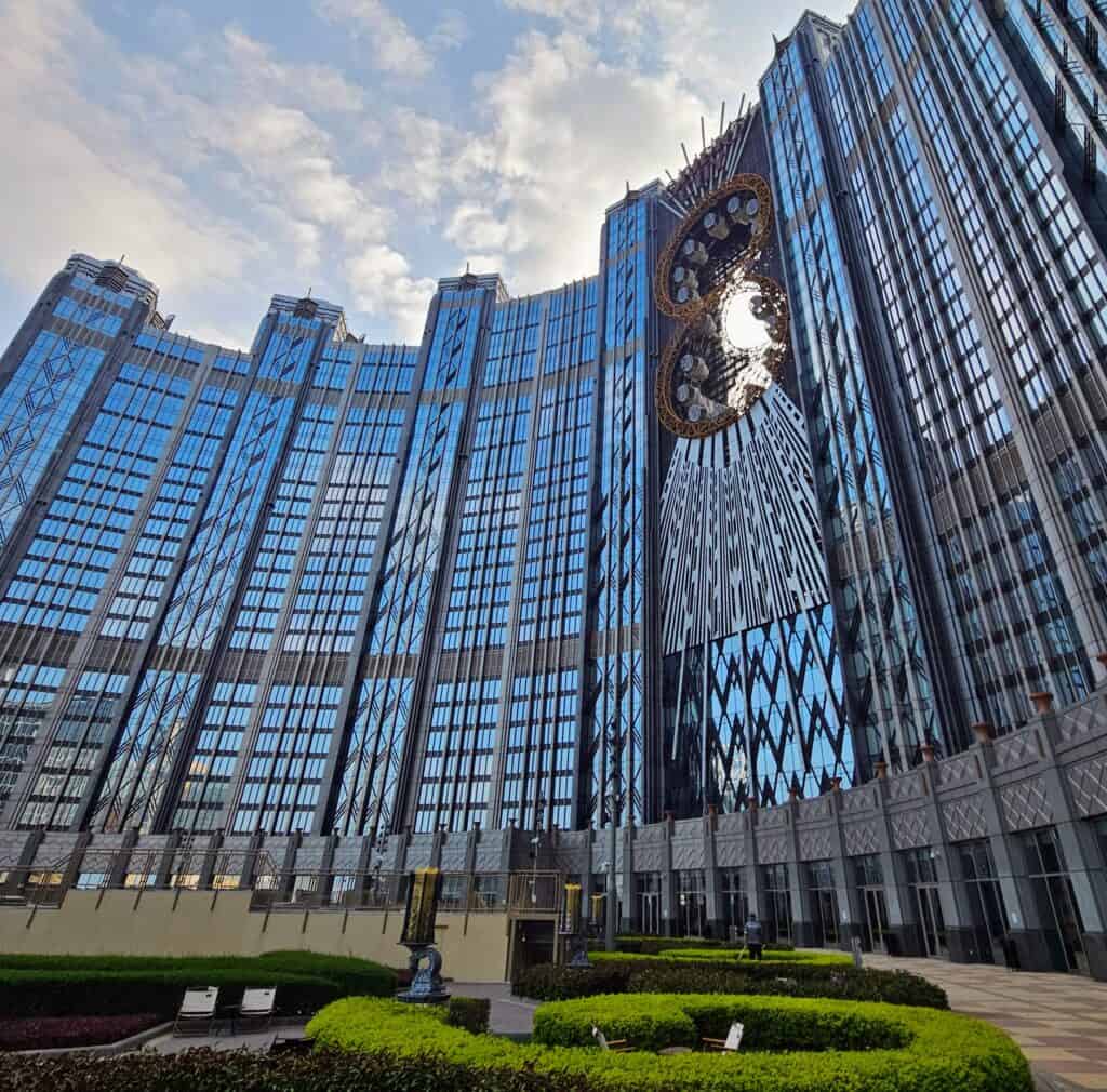 The towering Studio City hotel and casino in Macau with its futuristic glass facade and signature figure-eight-shaped golden structure bridging two towers. The building reflects the sky while neatly trimmed hedges and seating areas line the foreground.