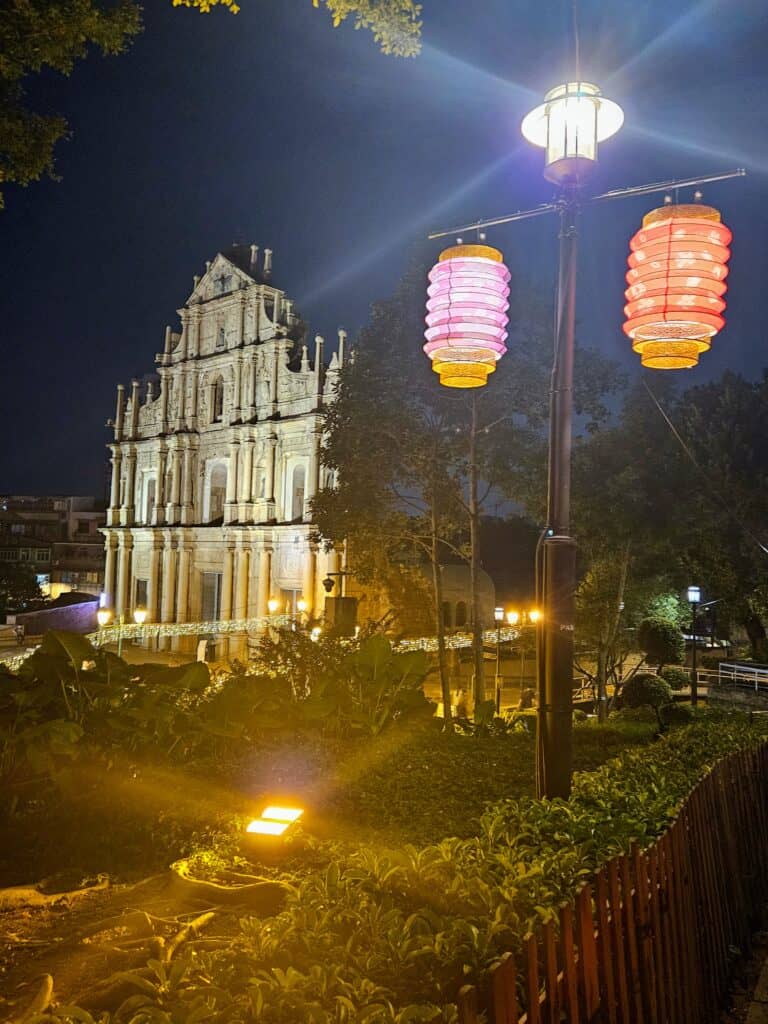 The Ruins of St. Paul’s in Macau illuminated at night, with its historic stone facade glowing under warm lights. Colorful lanterns hang from a nearby streetlamp, adding festive charm to the lush, garden-lined foreground.