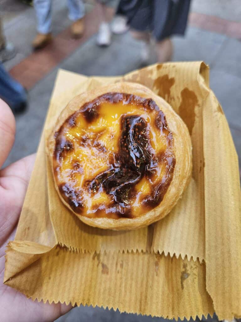 Close-up of a freshly baked Portuguese egg tart with a caramelized top and flaky golden crust, served on a brown paper wrapper. The tart is held in hand, with a blurred street scene and people's feet in the background.