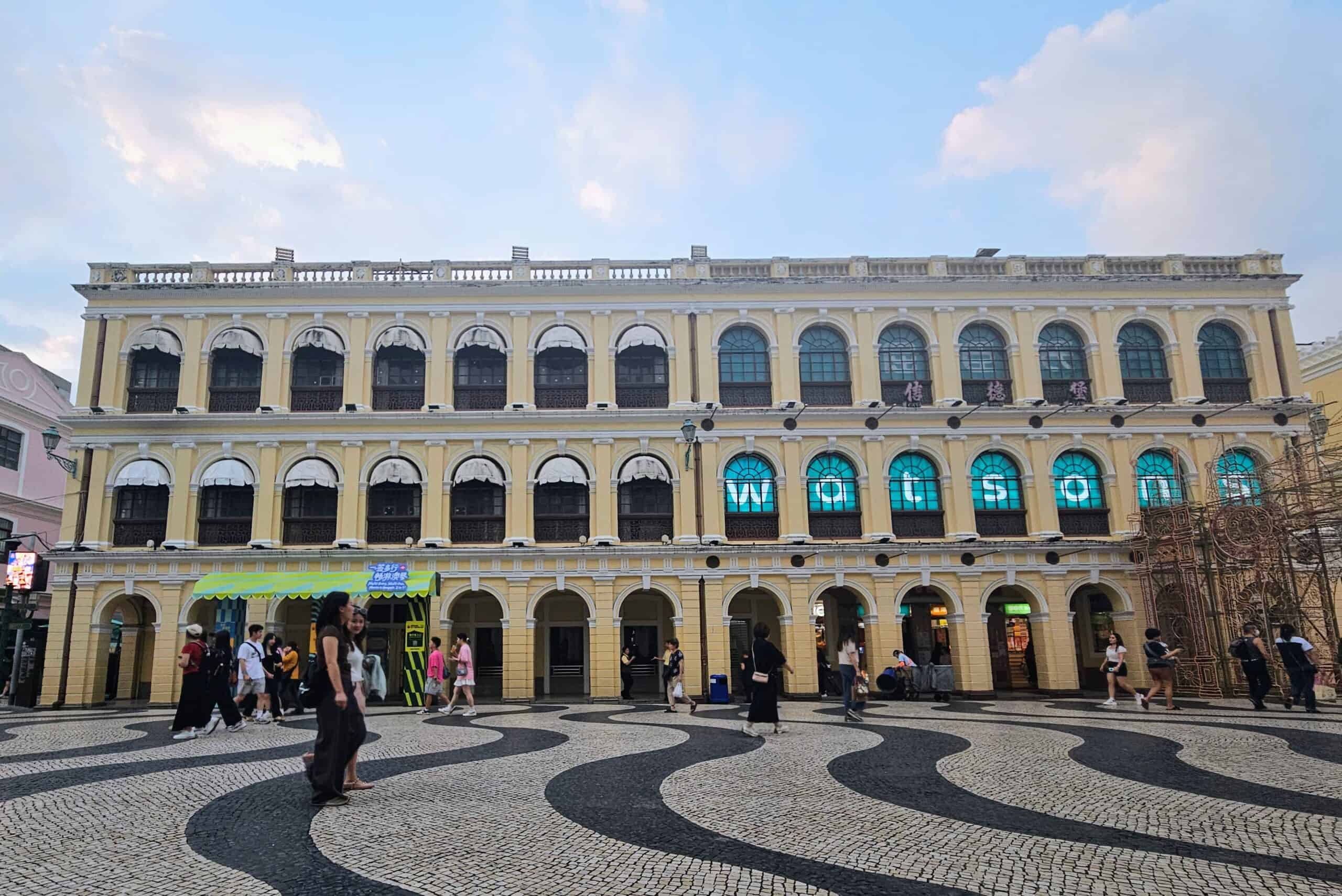 Wide-angle view of Senado Square in Macau at dusk, featuring a colonial-style yellow building with arched windows and patterned black-and-white mosaic pavement. Visitors stroll across the square, capturing the blend of Portuguese architecture and local culture.
