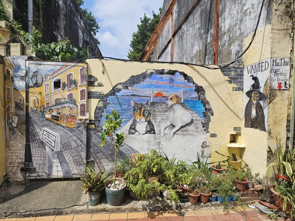 Colorful street art on a weathered wall in Macau depicts a tram scene, a seaside sunset with a cat and dog, and a witch-themed “WANTED” sign. The mural is framed by potted plants and captures the city’s playful, artistic charm in a hidden alleyway.