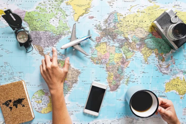 Flat lay of hands pointing at a world map surrounded by travel essentials including a compass, vintage camera, coffee mug, smartphone, journal, and a toy airplane. This image sets the tone for planning a multi-city trip, capturing the excitement of choosing destinations and organizing travel logistics.