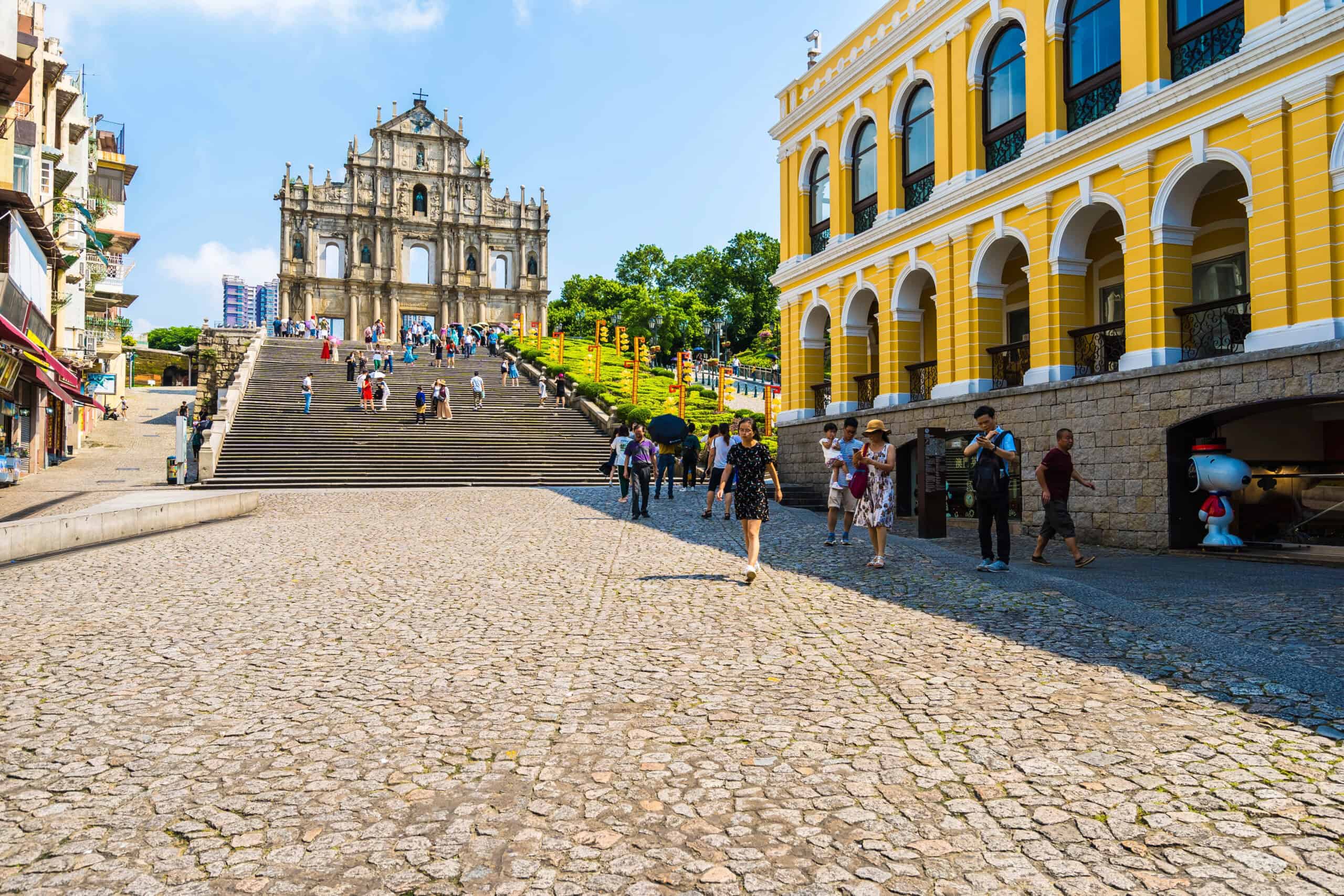 Tourists walk toward the iconic Ruins of St. Paul’s in Macau on a sunny day, with the stone facade of the 17th-century church rising above a wide staircase. Bright colonial-style buildings and cobblestone streets highlight Macau’s blend of Portuguese and Chinese heritage.