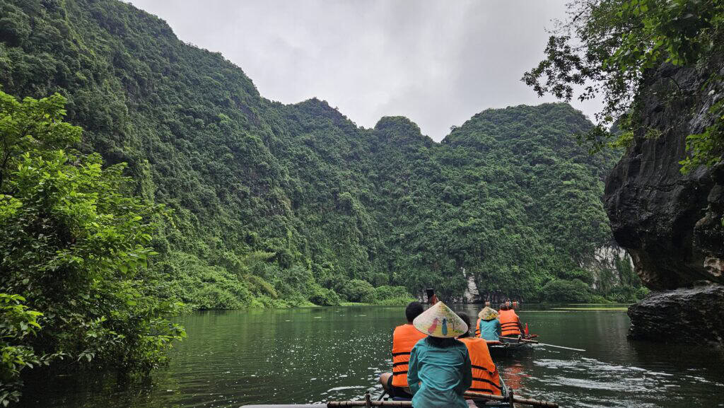 Tourists wearing life jackets ride in small rowboats through a lush green river valley surrounded by towering limestone cliffs in Ninh Binh, Vietnam. A guide in a conical hat paddles at the rear, showcasing a peaceful nature experience on a multi-city Southeast Asia itinerary.