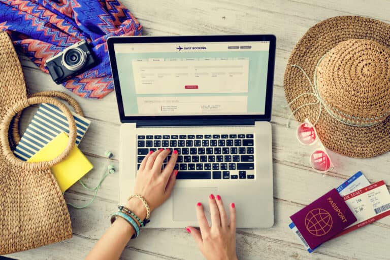 Overhead view of a person using a laptop on a wooden surface with a travel booking website open, surrounded by vacation essentials including a passport, airline tickets, sunglasses, a straw hat, a camera, and a woven beach bag. The scene captures the travel planning process for a multi-city trip.