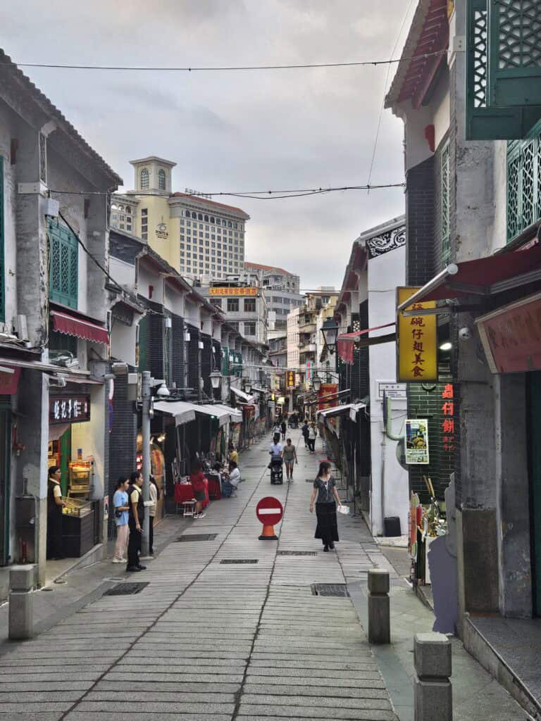 Lively pedestrian street in Macau lined with small shops, food stalls, and signs in Chinese characters, with people walking between historic low rise buildings.