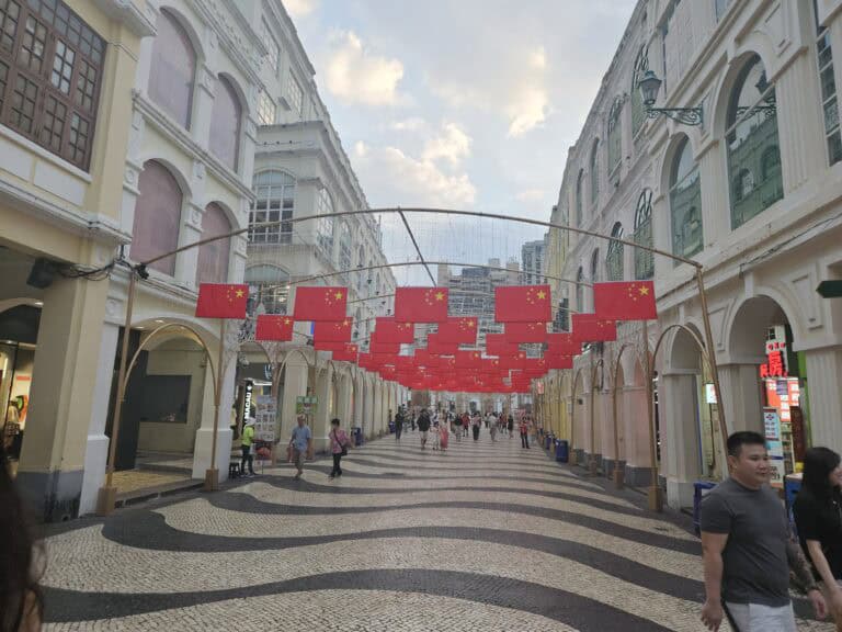 Senado Square in Macau with black and white wave patterned tiles and rows of red flags overhead, surrounded by pastel colonial style buildings and pedestrians.