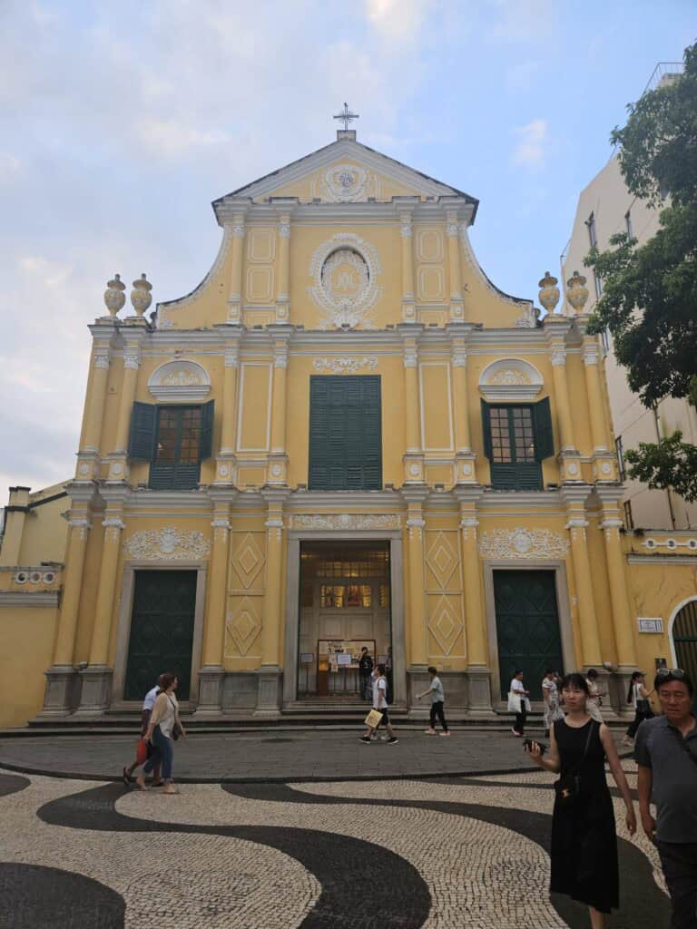 Yellow historic church in Macau with green shutters and ornate white detailing as people walk across the patterned square in front. The landmark is one of the cultural sights near BeLive and More hotel in Macau.