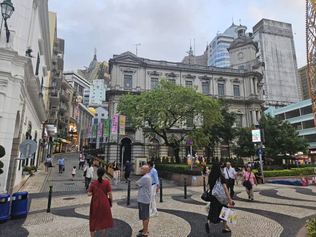 Busy Macau square with colonial style buildings, trees, and people walking across the patterned pavement, showing the central area near popular places to stay.