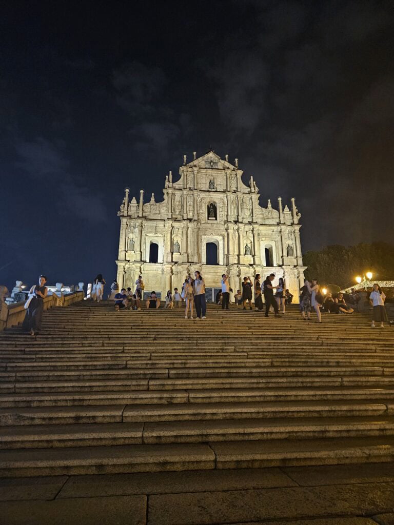 The Ruins of St Pauls in Macau lit up at night with people sitting and standing along the wide stone steps leading up to the historic facade. The scene shows the lively evening atmosphere near one of the most iconic landmarks close to many places to stay in Macau.