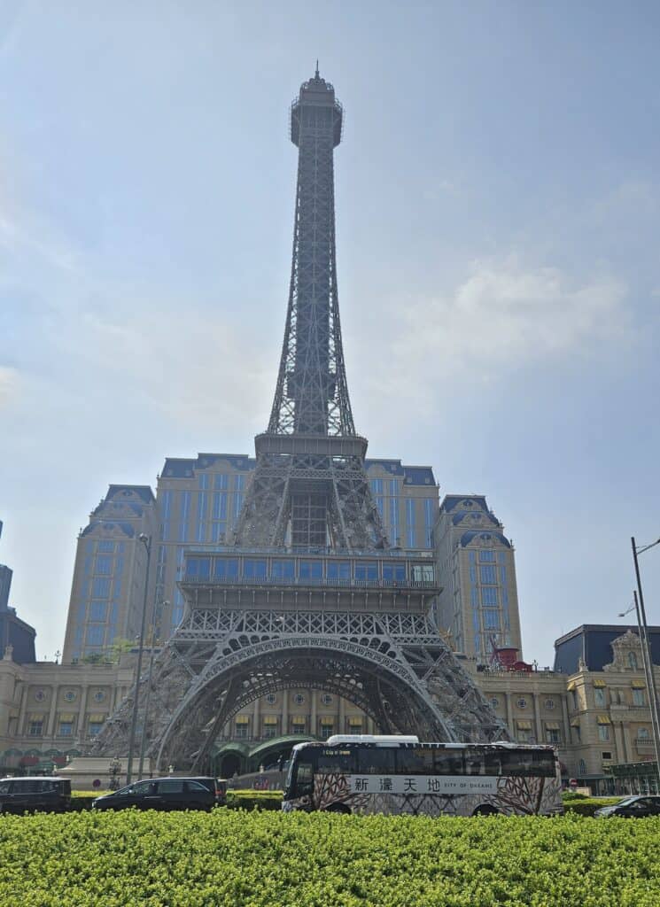 Eiffel Tower replica at The Parisian Macao rising above the Cotai Strip with resort buildings behind it and traffic passing in front. The scene highlights the Cotai area where many large resorts and hotels are located when choosing where to stay in Macau.