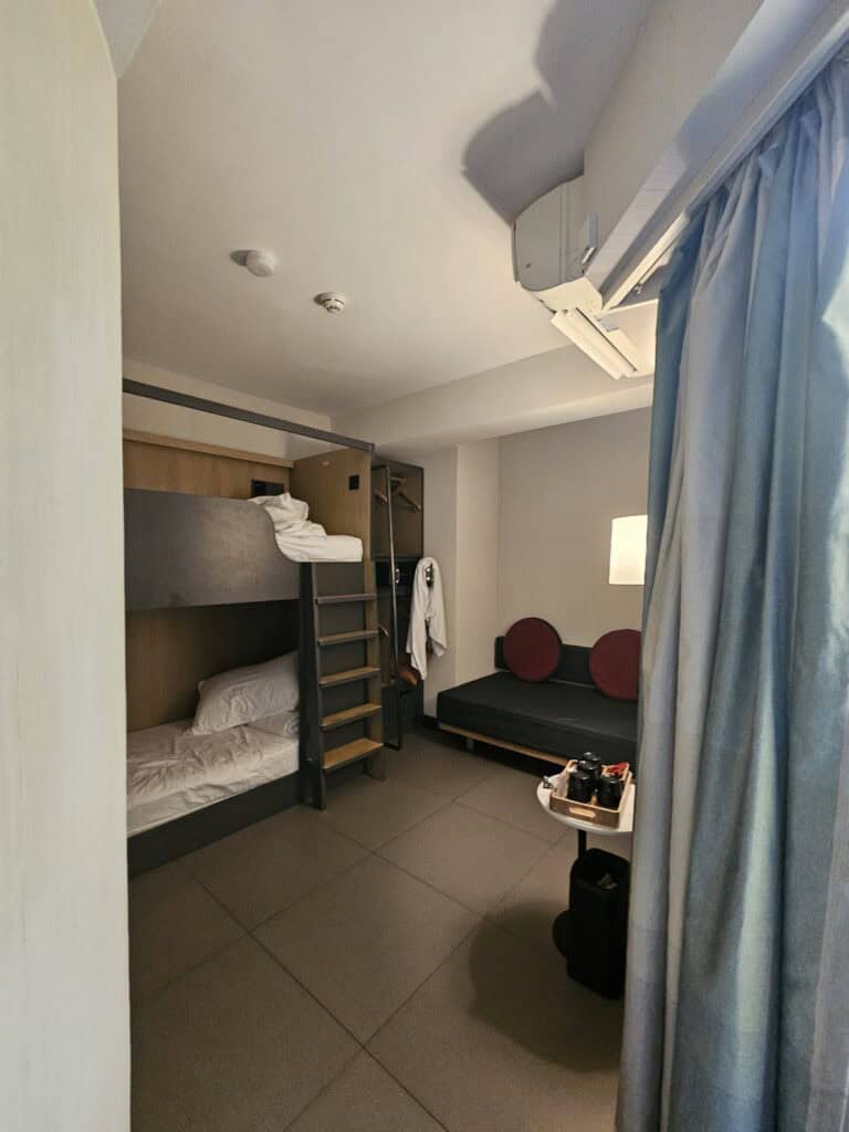 Full view of a BeLive and More hotel room in Macau with bunk beds, a small sofa, side table with tea set, and curtains covering the window. The compact layout gives a clear sense of space when deciding where to stay in Macau.