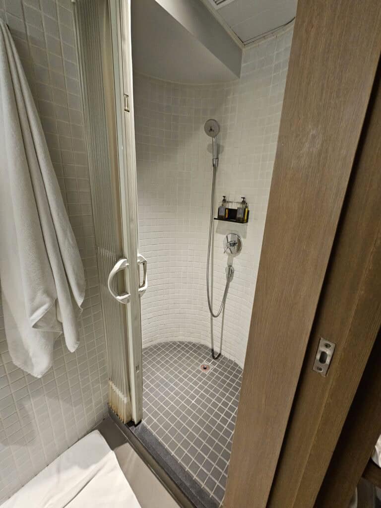 Small tiled shower area in a BeLive and More hotel bathroom with a handheld showerhead and toiletries mounted on the wall. The design reflects a compact but modern bathroom setup in Macau accommodation.