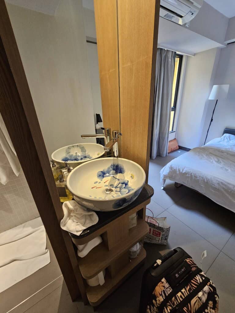 Open sink area in a BeLive and More hotel room with a decorative bowl basin, mirror, and view into the bedroom with luggage nearby. The layout shows how bathroom and sleeping spaces are integrated in a compact Macau stay.