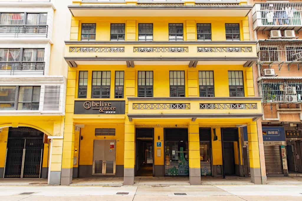 Bright yellow exterior of BeLive and More hotel in Macau with signage reading "belive & more" above the entrance. The building stands along a street of mixed older and modern structures, indicating its central location.