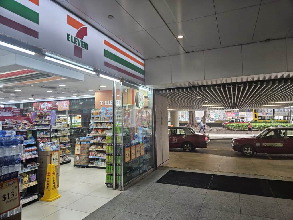 Entrance to a 7 Eleven convenience store in Hong Kong with shelves of snacks and drinks inside and taxis waiting outside on the street, where luggage can be stored before taking the Hong Kong to Macau ferry.