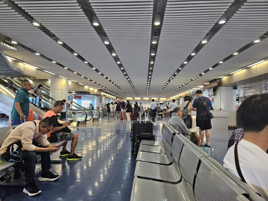 Wide view of a crowded ferry terminal in Hong Kong with passengers lining up near gates, others seated with suitcases, and escalators in the background before departure to Macau.