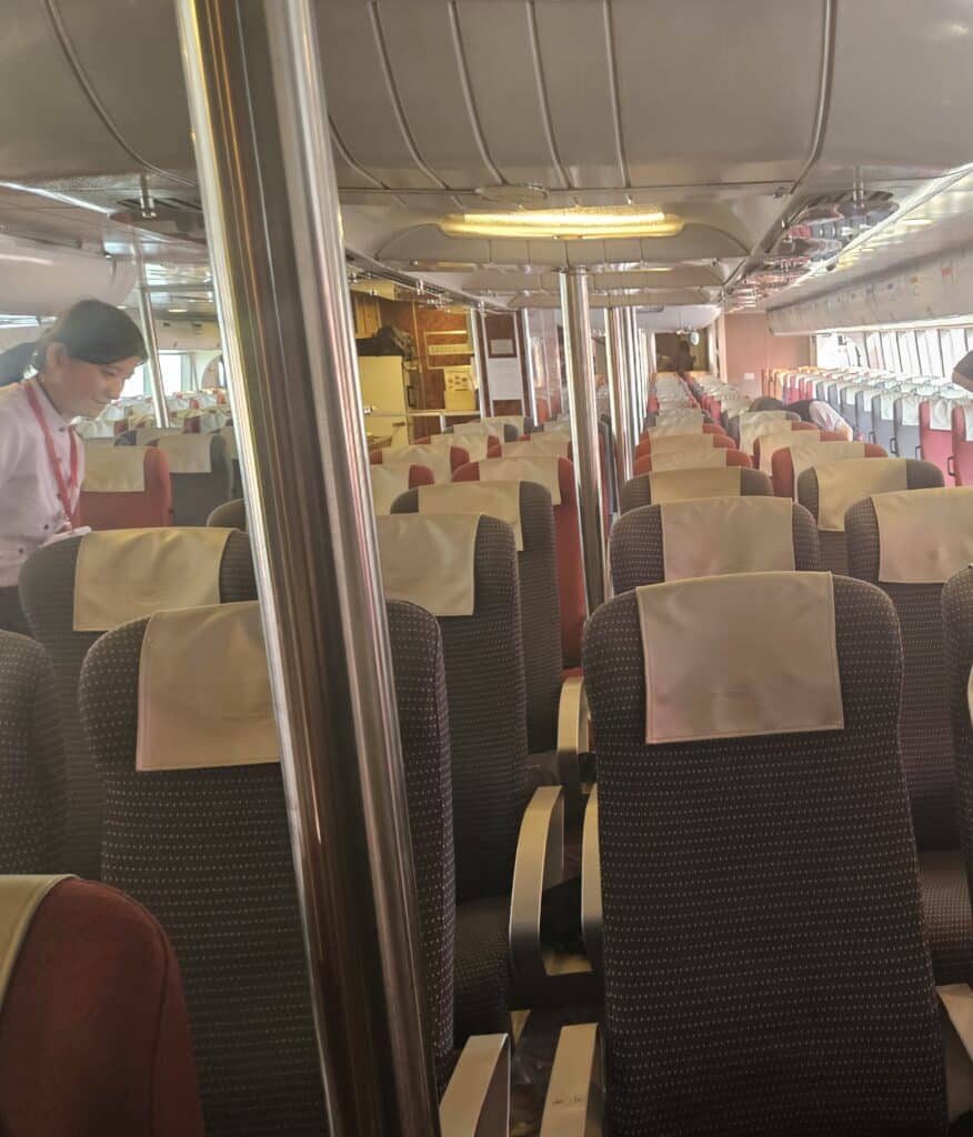 Rows of padded seats inside a TurboJET cabin with a crew member standing in the aisle and soft lighting overhead, showing the seating layout on the Hong Kong to Macau ferry before departure.