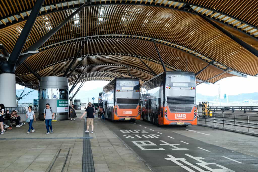 Double decker buses waiting at a covered bus stop at the Hong Kong Zhuhai Macau Bridge terminal with passengers nearby, showing the bridge bus as an alternative to the Hong Kong to Macau ferry.