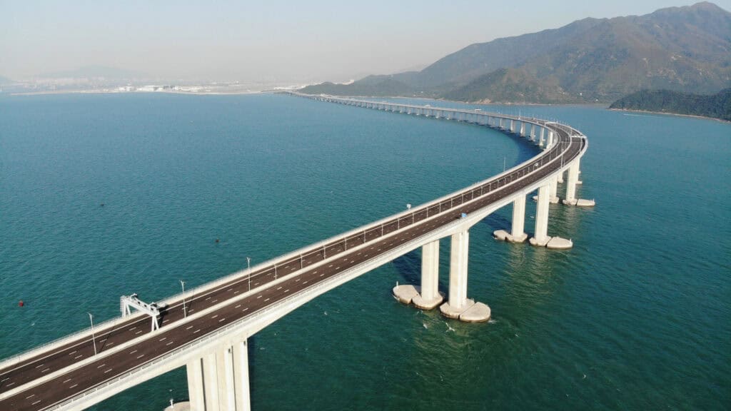 Aerial view of the Hong Kong Zhuhai Macau Bridge curving across blue water toward green mountains, showing the alternative route to the Hong Kong to Macau ferry.