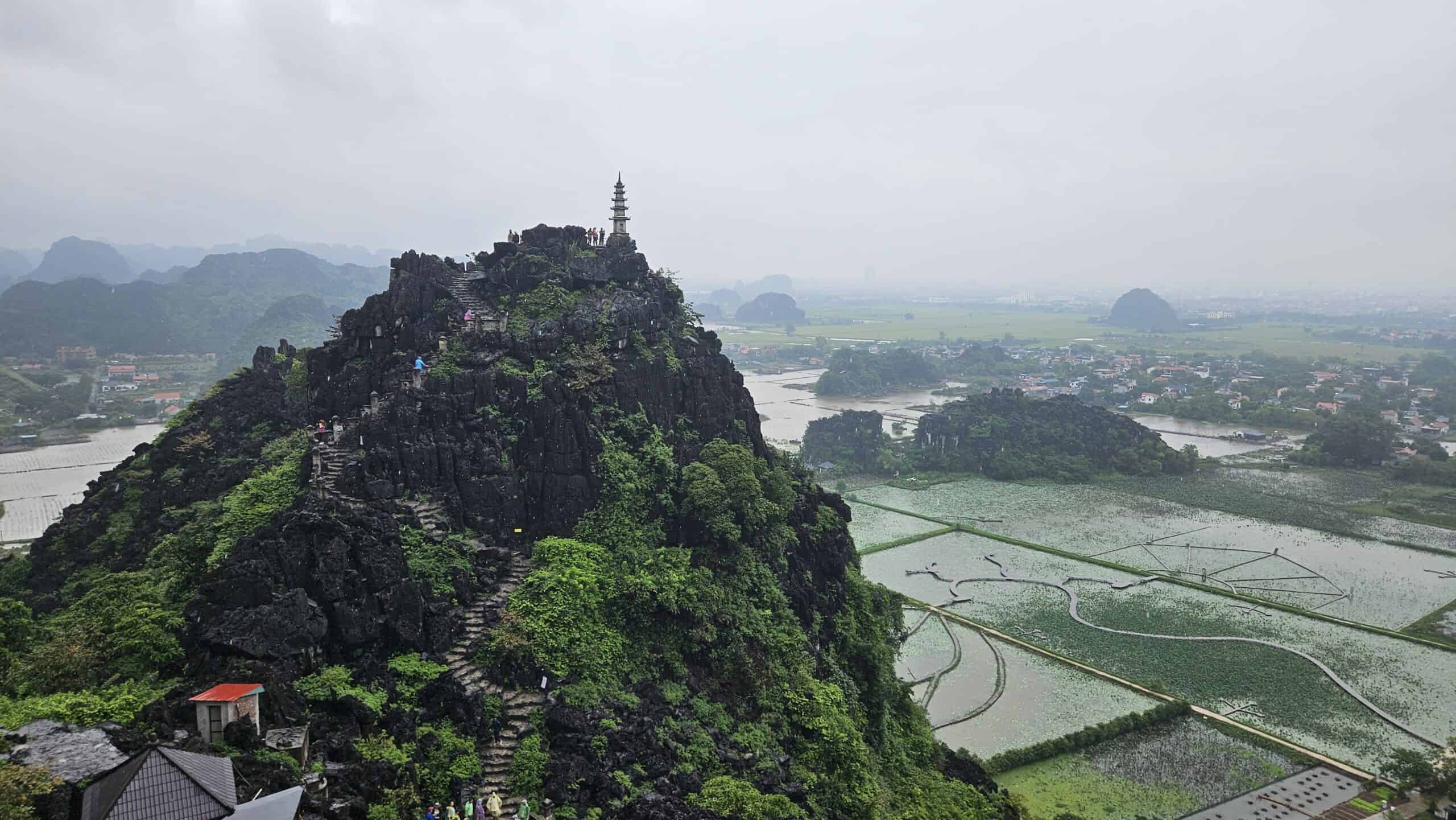 Panoramic view from Hang Mua viewpoint in Ninh Binh showing a rocky peak topped with a pagoda. Rice fields, winding waterways, and distant villages spread across the valley below.
