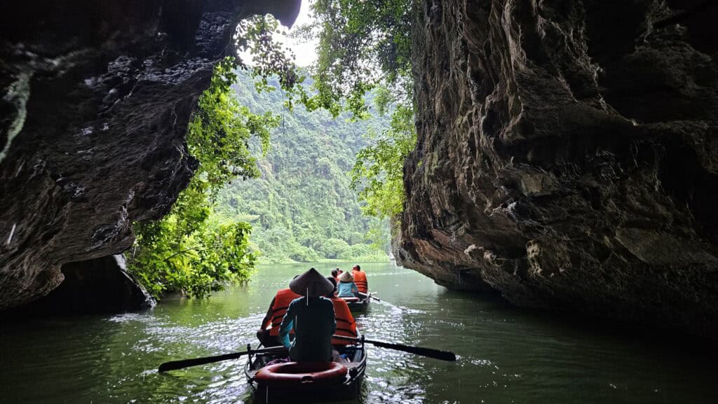 Small rowing boat passes through a cave opening in Ninh Binh surrounded by towering rock walls and hanging vines. Passengers wearing orange life jackets head toward the bright green river outside.