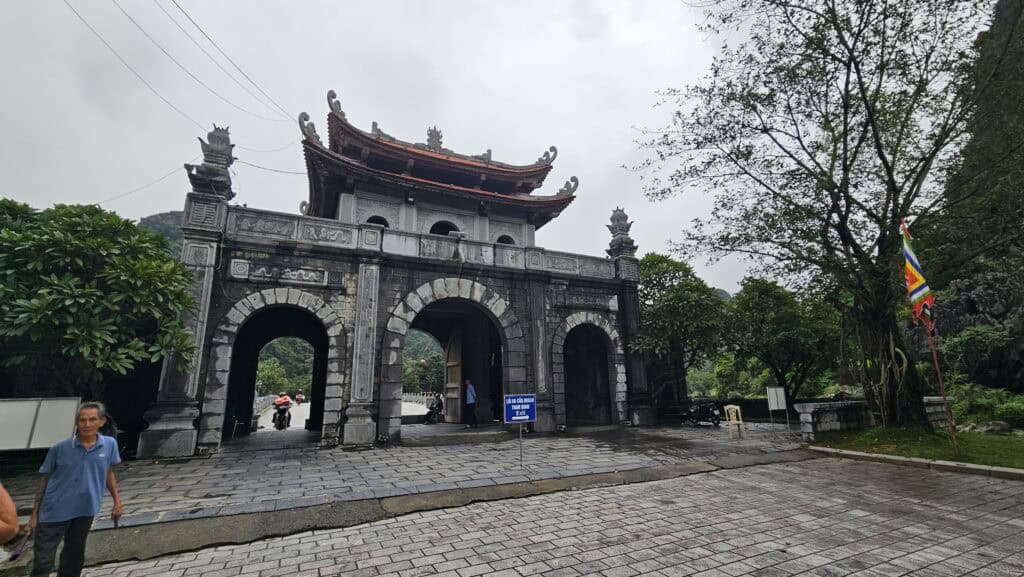 Large stone entrance gate with curved pagoda roof at a temple site in Ninh Binh. The rain soaked courtyard and cloudy sky frame the dramatic mountain landscape behind it.