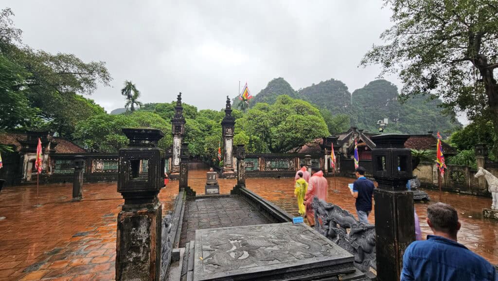 Historic temple courtyard in Ninh Binh with wet red brick paving after rain. Visitors in colorful ponchos walk between stone gates and flags with limestone mountains in the background.