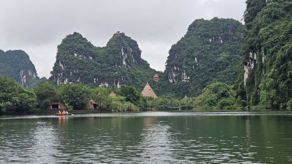 Rowboat glides across a calm green lake in Ninh Binh with towering limestone karsts rising behind it. Traditional thatched structures and misty mountains create a peaceful scene on this day trip from Hanoi.