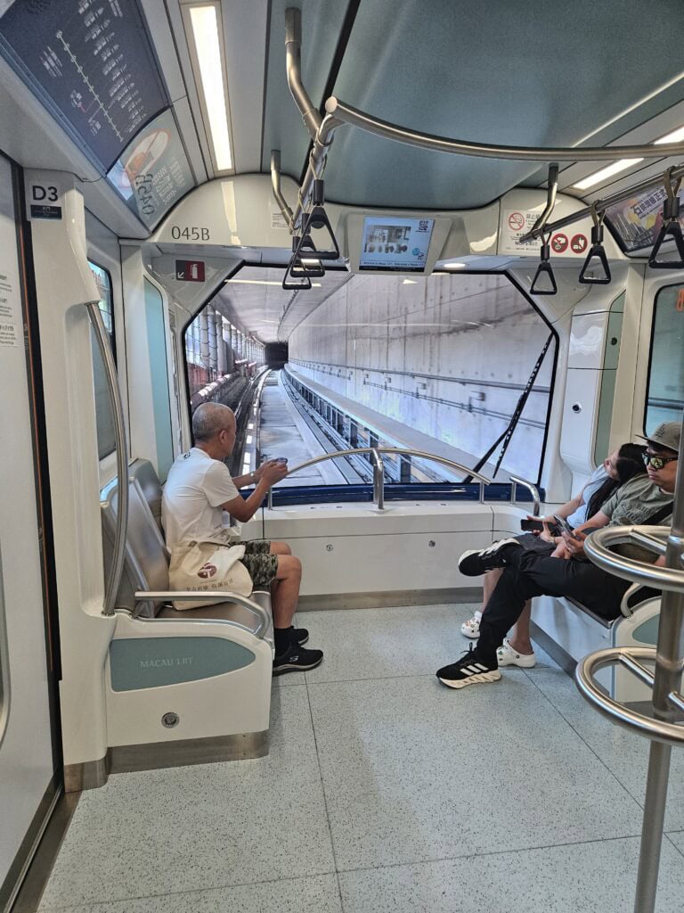 Interior of the Macau LRT looking out through the front window into a concrete tunnel while a few passengers sit facing each other. The car number "045B" is visible above the window and the scene shows what riding the light rail feels like for anyone researching how to get around Macau.