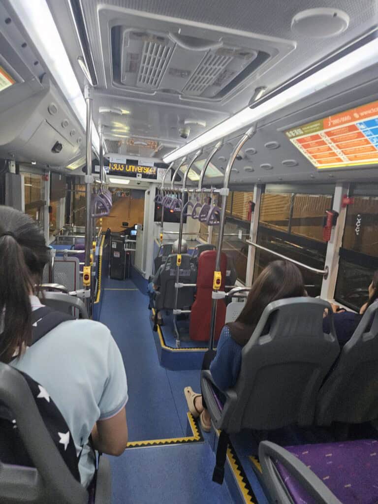 Inside a Macau public bus at night with passengers seated on both sides and purple hand straps hanging above the aisle. The route display at the front glows with partially visible text that appears to read "universi" which helps show what taking the bus is like when figuring out how to get around Macau.