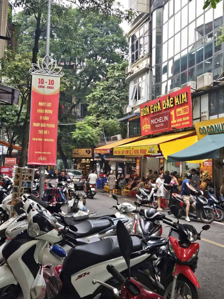 Busy Hanoi street filled with parked motorbikes, people dining outside, and a bright red sign reading "Bún Chả Đắc Kim MICHELIN Selected Since 1966," capturing the lively atmosphere when choosing where to stay in Hanoi.