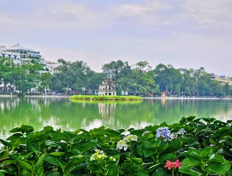 Hoan Kiem Lake in Hanoi with the Turtle Tower centered on a small island, framed by green plants and flowers in the foreground, highlighting a key landmark near where to stay in Hanoi.