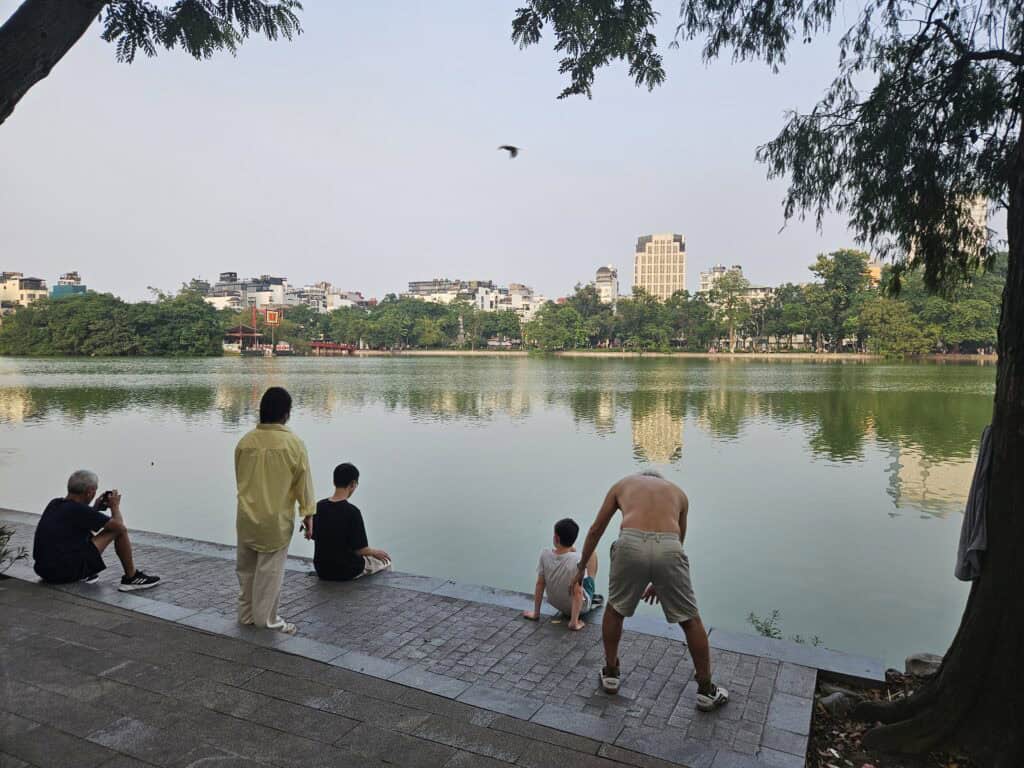 Locals relaxing by Hoan Kiem Lake in Hanoi with calm water, city buildings in the background, and people sitting along the edge, showing the peaceful side of where to stay in Hanoi.