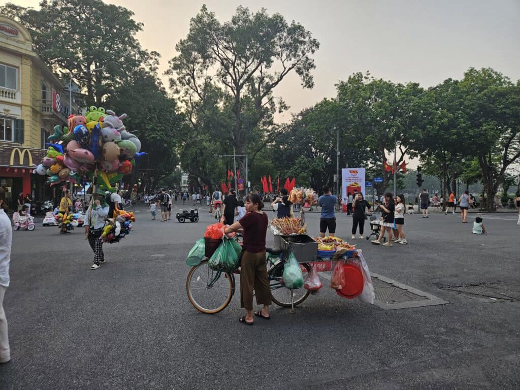 Street vendor in Hanoi standing beside a bicycle loaded with snacks and goods, with pedestrians and a McDonalds sign in the background, showing everyday life when exploring where to stay in Hanoi.