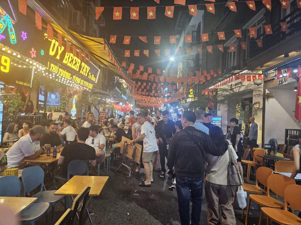 Crowded Hanoi night street lined with restaurants and glowing signs including "Uy Beer Tây," with red flags overhead and people dining outdoors, highlighting nightlife near where to stay in Hanoi.