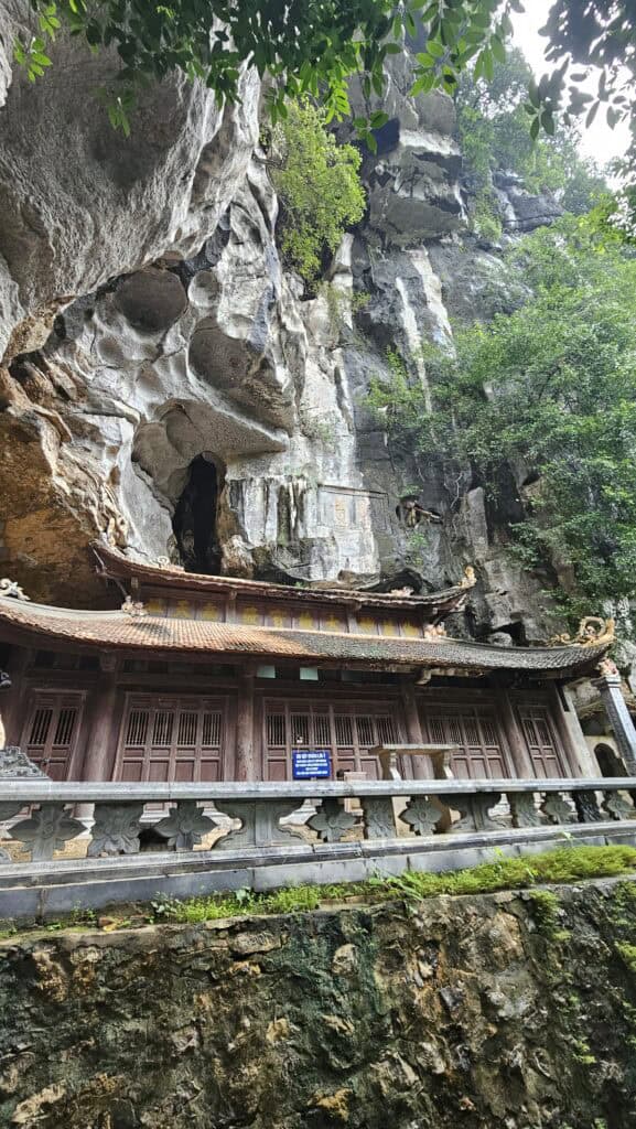 Ancient temple built into the base of a towering limestone cliff in Ninh Binh. Weathered wooden doors and carved stone details sit beneath cave openings and hanging greenery.