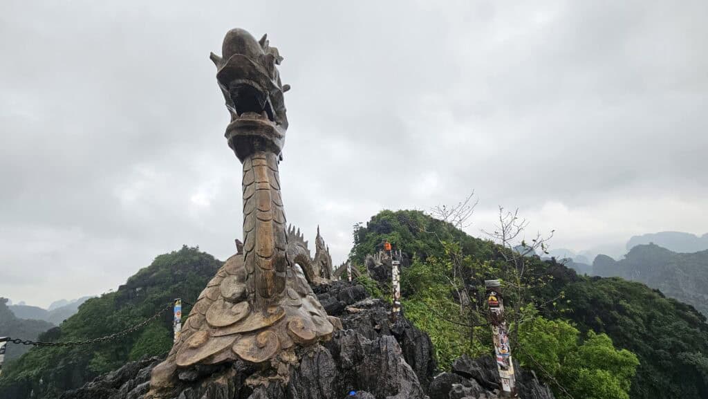 Large carved dragon sculpture stands on a rocky summit in Ninh Binh at Mua Caves under a cloudy sky. The dramatic viewpoint overlooks layers of green karst mountains.