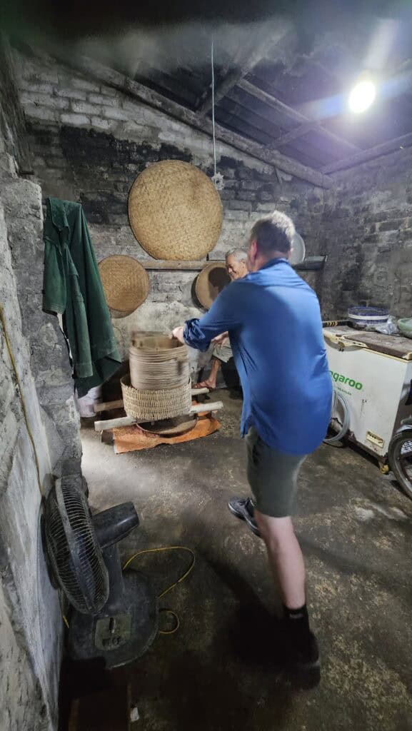 Traveler learns how rice wine is made inside a rustic workshop in Ninh Binh. Woven bamboo trays hang on the wall while an older local guide demonstrates the traditional spinning process used during