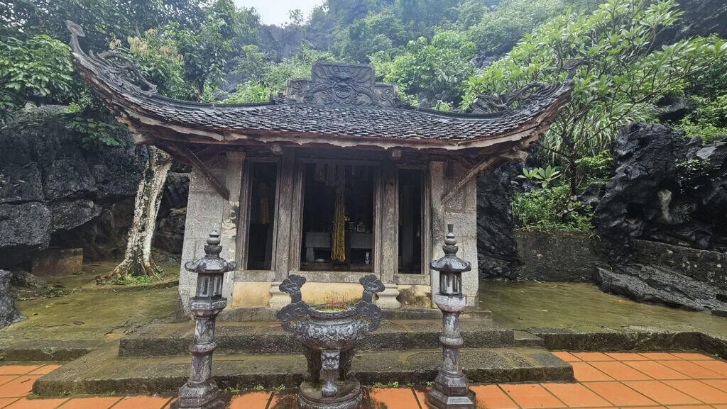 Small stone shrine surrounded by dramatic black rock formations and tropical plants in Ninh Binh. Two lantern pillars and an incense burner stand in front of the rustic temple building.