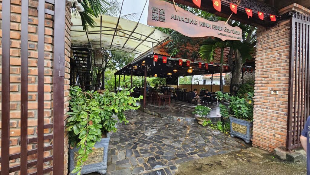 Entrance to Amazing Ninh Binh Garden restaurant with brick pillars, tropical plants, and covered outdoor seating. Red flags hang above the stone walkway on a rainy day.