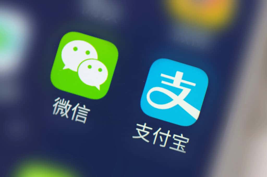 Close up of two phone app icons on a dark screen. The green WeChat icon is labeled "微信" and the blue Alipay icon is labeled "支付宝" showing common payment apps travelers may use while getting around Macau.