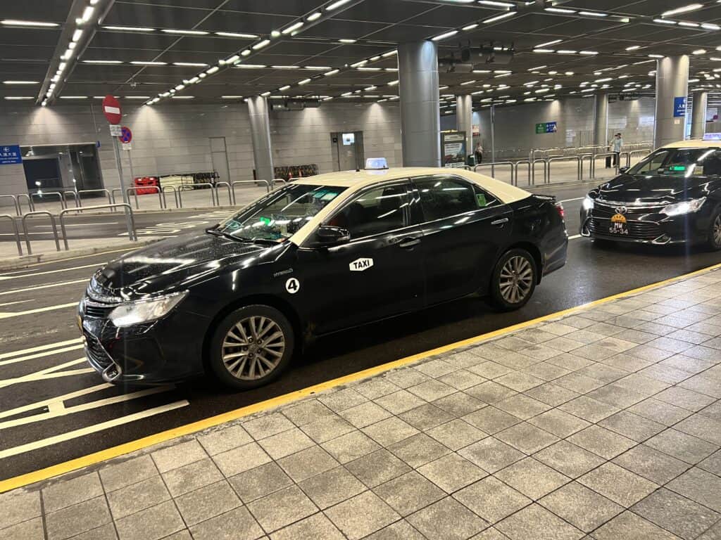 Black taxi with a cream roof waiting at a curbside pickup area under bright ceiling lights. This shows the taxi option for how to get around Macau especially when arriving at a terminal or needing a direct ride.