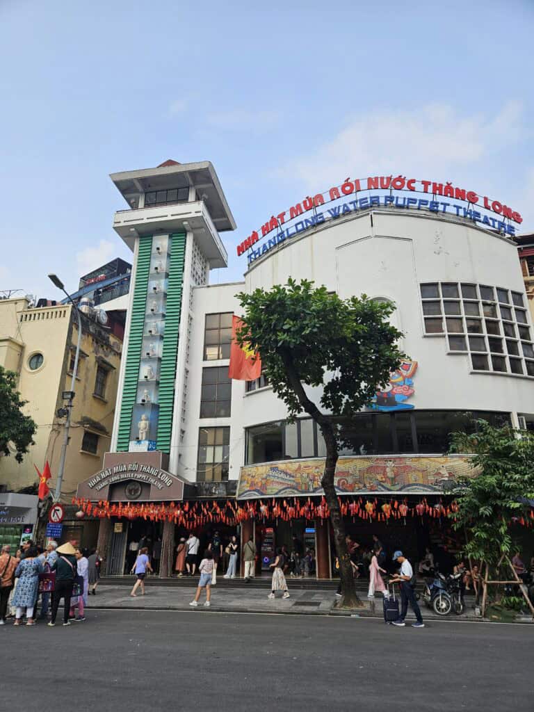 Exterior of Thang Long Water Puppet Theatre in Hanoi with curved white facade and large rooftop sign reading "Thang Long Water Puppet Theatre." People gather outside the entrance beneath red flags while trees line the street.