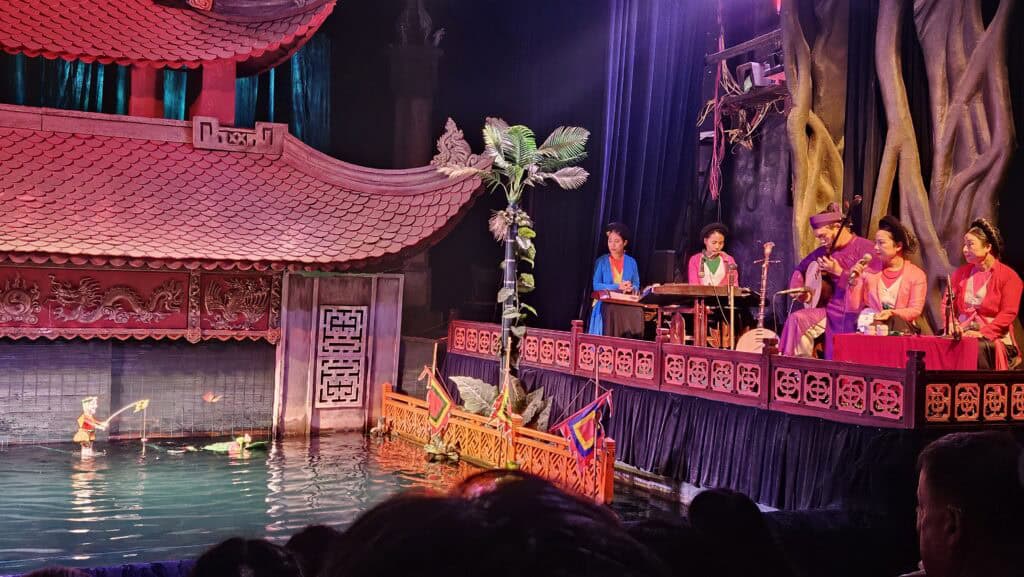 Musician in traditional dress plays a string instrument beside a drummer on the side stage at Thang Long Water Puppet Theatre. Large tree root scenery and blue lighting frame the live music that accompanies the Thang Long water theatre puppet show.