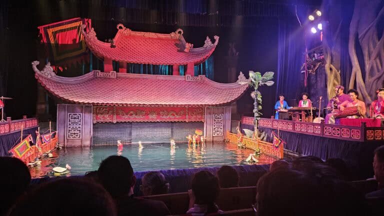 Wide view of the Thang Long Water Puppet Theatre stage with several colorful puppets rising from the water in front of a red temple set. Musicians perform live at the side as the audience watches the Thang Long water theatre puppet show in Hanoi.